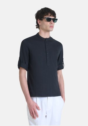 Camisa henley negra de manga corta con un escote curvado y tapeta de botones, cuenta con puños enrollados y un sutil logo en el pecho.