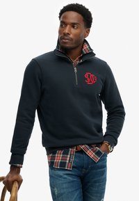 Maglione blu navy con collo a zip e logo rosso, indossato sopra una camicia a quadri. Caratterizzato da una texture morbida e una vestibilità casual, abbinato a jeans in denim.