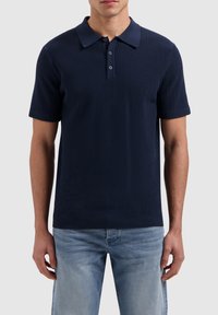 Marineblauwe polo met een textuurgebreid ontwerp, korte mouwen en een kraag. Twee knopen bij de hals en een ribgebreide zoom. Draagt samen met lichte jeans.