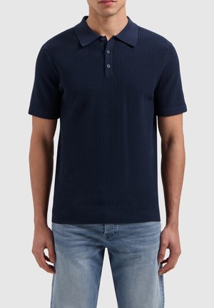 Marineblauwe polo met een textuurgebreid ontwerp, korte mouwen en een kraag. Twee knopen bij de hals en een ribgebreide zoom. Draagt samen met lichte jeans.