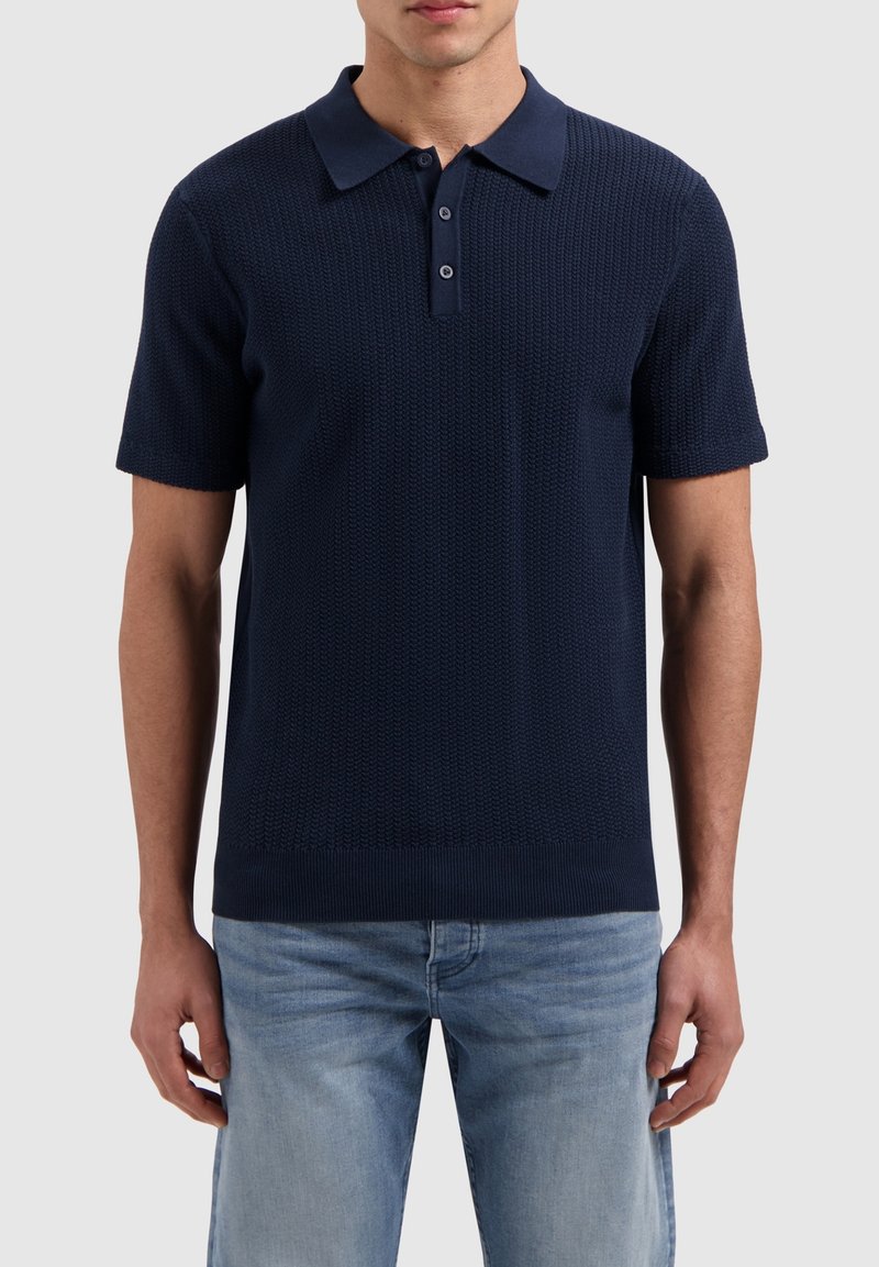 Marineblauwe polo met een textuurgebreid ontwerp, korte mouwen en een kraag. Twee knopen bij de hals en een ribgebreide zoom. Draagt samen met lichte jeans.