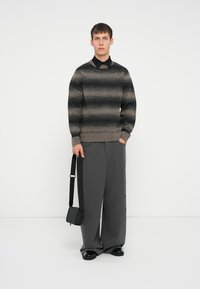 Men's striped sweater in dark grey and beige with a crew neck, paired with wide, grey pinstripe trousers and a black crossbody bag.