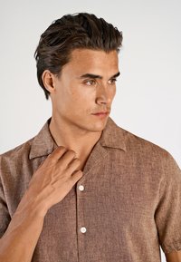 Bruin linnen overhemd met korte mouwen en een gestructureerde stof, een knoopsluiting aan de voorkant en een brede kraag. Model past de kraag aan met één hand.