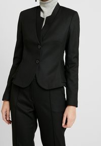 Zwarte op maat gemaakte blazer met een gestructureerd ontwerp, reverskraag, twee knopen en textiel met een textuur. Gecombineerd met bijpassende zwarte broek.