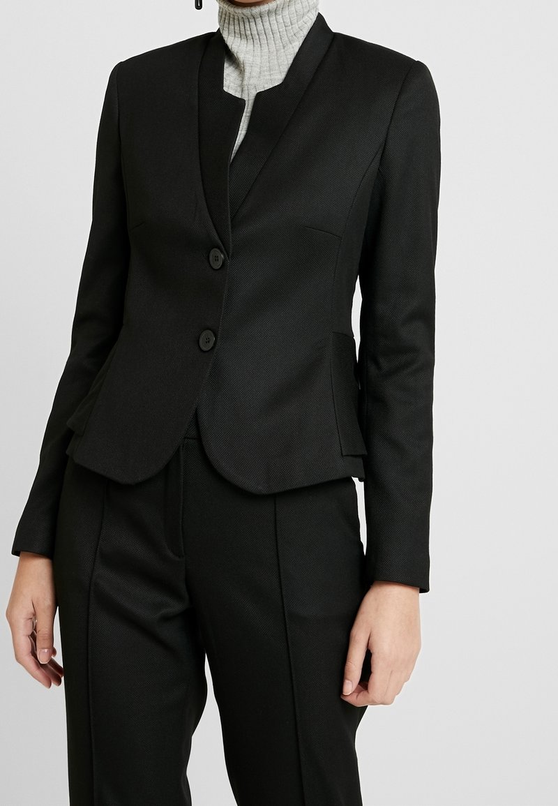Zwarte op maat gemaakte blazer met een gestructureerd ontwerp, reverskraag, twee knopen en textiel met een textuur. Gecombineerd met bijpassende zwarte broek.