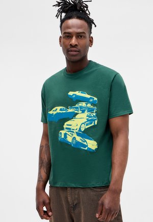 Άνδρας με dreadlocks φοράει ένα πράσινο μπλουζάκι με ένα στοίβαγμα γραφικών έξι κίτρινων και μπλε vintage και μοντέρνων αυτοκινήτων.