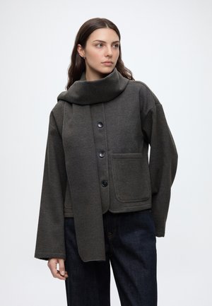 Kvinde med langt brunt hår iført en mørkegrå knappet jakke med stort tørklæde og mørkeblå jeans, kigger til siden.