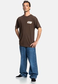 Brown cotton t-shirt with a rounded neckline and a logo patch on the front, paired with loose blue denim jeans and black slip-on shoes.