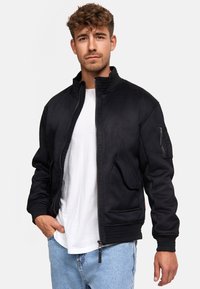 Chaqueta bomber negra con cuello alto, cremallera frontal y bolsillos laterales. Presenta puños de canalé y un bolsillo con cremallera en la manga. Llevada sobre una camiseta blanca.