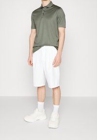 Man som bär en olivgrön kortärmad polo, vita knähöga shorts, vita strumpor och vita sneakers, stående mot en neutral bakgrund.