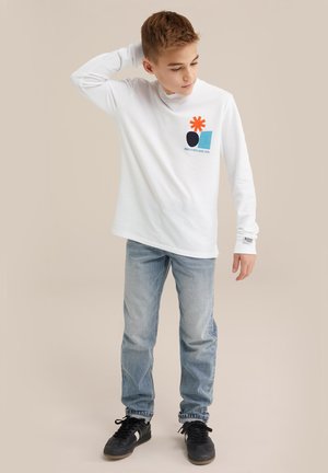 Langarmweißes T-Shirt mit einem bunten Grafikdesign, das einen roten Stern, ein blaues Quadrat und einen schwarzen Kreis zeigt; kombiniert mit hellblauen Jeans und schwarzen Sneakern.