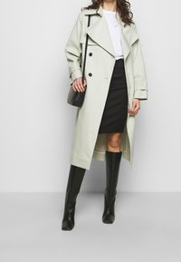 Trench coat verde claro con diseño de doble botonadura, grandes solapas y herrajes negros, llevado sobre una falda negra y botas negras hasta la rodilla.