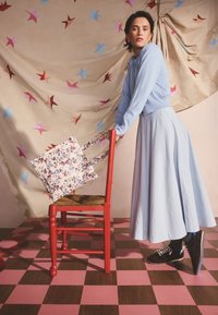 Moteris šviesiai mėlynos kardiganės ir sijono, atsirėmusi į raudoną kėdę su gėlėtu rankinės krepšiu, stovi ant rožinės ir rudos languotos grindų, fone žvaigždžių rašto fonas.