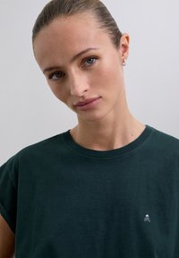 Dark green cotton T-shirt with short sleeves and a crew neck. Features a small skull and crossbones embroidery on the left chest area.