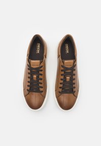 Zapatillas de cuero marrón con un acabado suave, cordones negros y suelas de goma blanca. El forro interior es de tela oscura con el logo.