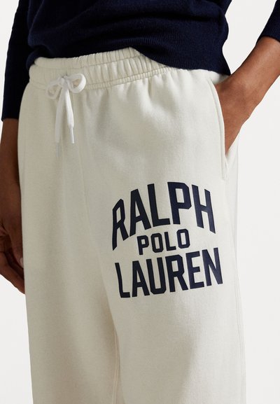 Cream-colored sweatpants featuring a prominent dark navy logo that reads "RALPH POLO LAUREN" on the left thigh. Elastic waistband and pockets.