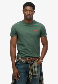 Mand iført grøn t-shirt med lille rød logo, blå jeans, brun bælte, ur og ternet skjorte knyttet om taljen på hvid baggrund.