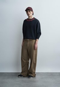 Άνδρας που φοράει μπορντό καπέλο, μαύρο φούτερ με κόκκινο κείμενο, φαρδιά καφέ παντελόνια και μαύρα παπούτσια στέκεται μπροστά σε έναν απλό γκρι τοίχο.