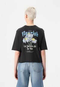 Juodas trumpas marškinėlis su gėlių grafika ir užrašu "Flourish". Trumpomis rankovėmis ir laisvu siluetu, pagamintas iš minkštos, tekstūruotos medžiagos.