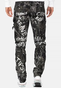 Pantalons cargo noirs avec éclaboussures de peinture blanche et texte, conçus avec des poches et une coupe fuselée. Les chaussures sont blanches avec des accents rouges et bleus.
