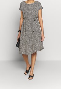 Vestido de patrones en blanco y negro con mangas cortas, cuello redondo, cintura ajustada, detalles de botones y una falda fluida. Combinado con zapatos negros de punta abierta.