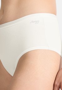 Gros plan sur une personne portant des slips en coton blancs avec la marque "Sloggi" sur la ceinture, montrant la hanche et la partie supérieure de la cuisse.