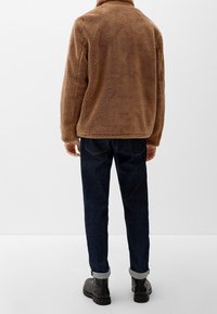 Person iført en brun fleecejakke, mørkeblå opprullede jeans og sorte støvler, stående med ryggen til mod en hvid baggrund.