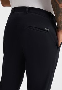 Schwarze Hose aus glattem Stoff mit schmalem Schnitt, Gürtelschlaufen und zwei Gesäßtaschen, von denen eine mit einem metallischen Logoakzent hervorgehoben ist.