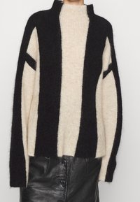 Femme portant un pull en peluche à rayures verticales noires et beiges, associé à une jupe en cuir noire boutonnée.