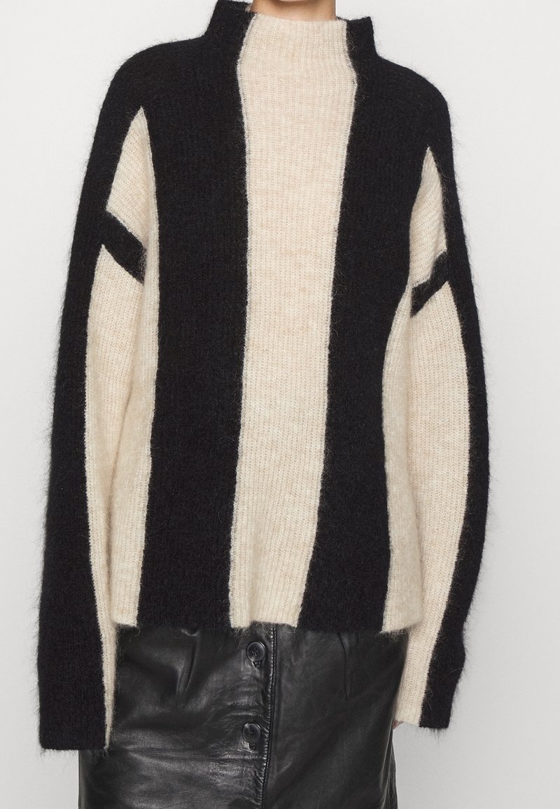 Femme portant un pull en peluche à rayures verticales noires et beiges, associé à une jupe en cuir noire boutonnée.