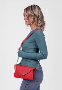 Červená kožená kabelka s překlopným víkem a zlatým akcentem, držena na řetízkové šňůře. Nosená přes tělo na šedém pozadí.