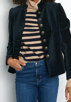 Femme portant une veste en velours bleu marine avec des boutons en laiton sur un pull col roulé beige et bleu marine à rayures et un jean bleu, main dans la poche.