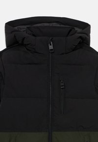 Veste rembourrée noire et verte avec un col haut, fermeture éclair à l'avant et une poche zippée sur le côté gauche. Texture lisse, design minimal.