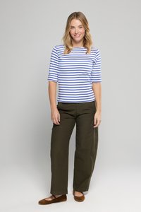 Kvinde, der smiler, iført en blåt og hvidt stribet kortærmet bluse, olivengrønne brede bukser og brune flade sko, stående mod en ensfarvet baggrund.