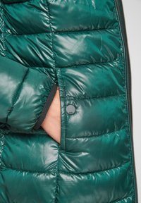 Teal oppustelig jakke med en quilted design og glansfuld finish, har en sidelomme med en trykknap og sort indvendig kant.