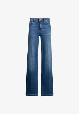 Femme portant un haut blanc sans manches et un jean bleu taille haute à jambes larges, avec des chaussures blanches pointues, debout avec une main dans la poche.