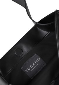 Intérieur d'un sac à main en cuir noir avec une texture lisse, une étiquette portant la mention "TUCANO MILANO 1985" et un détail de fermeture à pression circulaire.