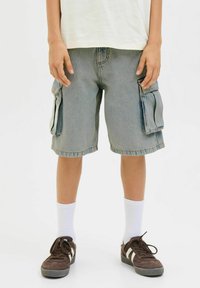 Shorts cargo en denim bleu clair avec deux poches latérales, bords effilochés et coupe décontractée, portés avec des chaussettes blanches et des baskets marron.