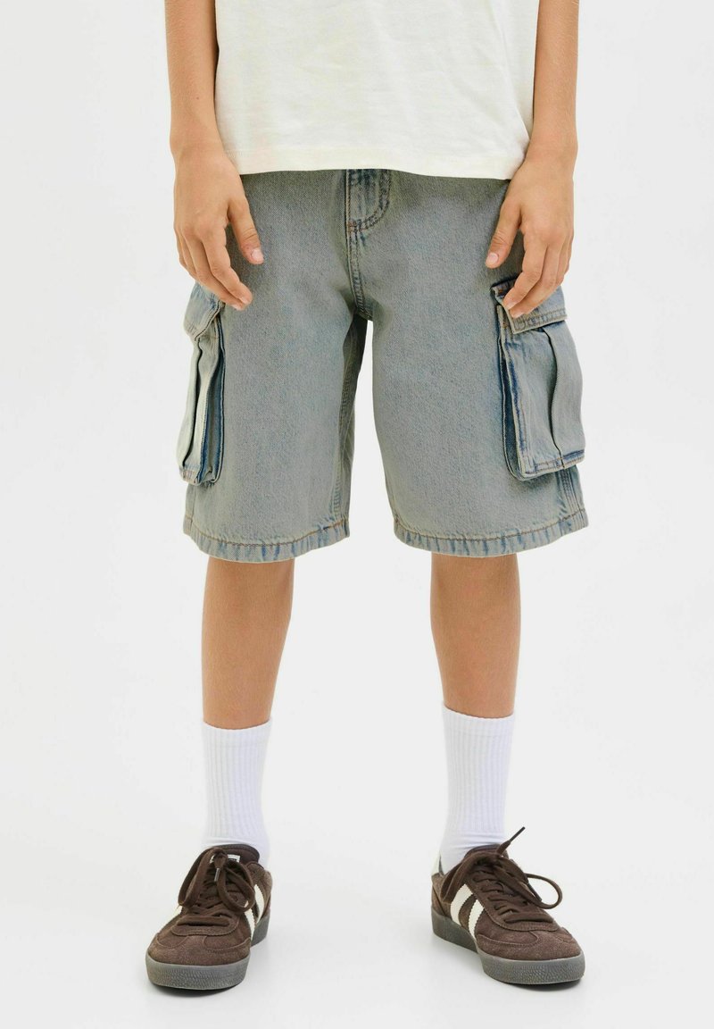 Shorts cargo en denim bleu clair avec deux poches latérales, bords effilochés et coupe décontractée, portés avec des chaussettes blanches et des baskets marron.