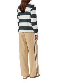 Vrouw in beige wijde broek met zwart-witte zijkanten en een groen-wit gestreept shirt met lange mouwen, staand met haar rug naar ons toe.