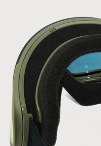 Zbliżenie na oliwkowozielone gogle narciarskie z grubym czarnym piankowym wyściełaniem i odbijającą się niebieską krawędzią soczewki, na białym tle.