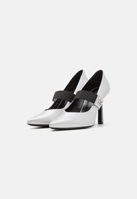 Pantofi cu toc înalt argintii, cu vârf ascuțit, bandă elastică neagră peste glezna piciorului și un design elegant, cu suprafețe strălucitoare și toc subțire.