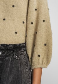 Pull en tricot beige avec des pois noirs, présentant des manches bouffantes amples. Associé à un pantalon en denim noir taille haute avec une fermeture à bouton.