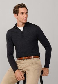 Donkergrijze, lange mouwen zip-up trui met een gladde textuur, gecombineerd met licht kaki broek en een leren bruine riem, zittend op een kruk.