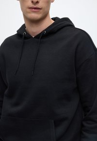 Crna pamučna dukserica s kapuljačom s prednjim džepom, podesivim vezicama i opuštenim krojem. Mekana tekstura i minimalistički dizajn.