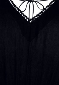 Svart klänning med djup V-ringning och scalloped kant, lättviktsmaterial och delikata band som knyts i ryggen. Texturerad ytdetalj synlig.