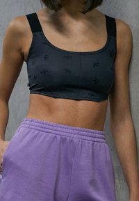 Černý sportovní crop top s hladkou texturou, zdobený černými logy, širokými ramínky a přiléhavým střihem, kombinovaný s fialovými tepláky.