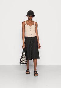 Top de linho bege claro com alças ajustáveis e botões à frente, combinado com uma saia midi preta com botões. Mala tecida em preto e branco e sandálias.