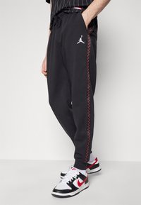Černé joggingové kalhoty s elastickým pasem na šňůrku, s červeným bočním prošitím a bílým logem Jumpman na levém boku. V kombinaci s černými a bílými teniskami.