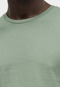 Gros plan d'une personne portant un t-shirt vert clair uni à col rond, montrant le cou et le haut de la poitrine.